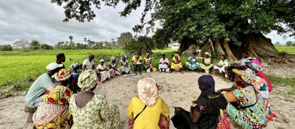 Members of a women's savings group sit in a circle beneath a tree.
