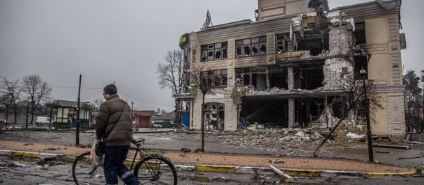 A man walks with a bicycle past a heavily damaged building on a gloomy street.