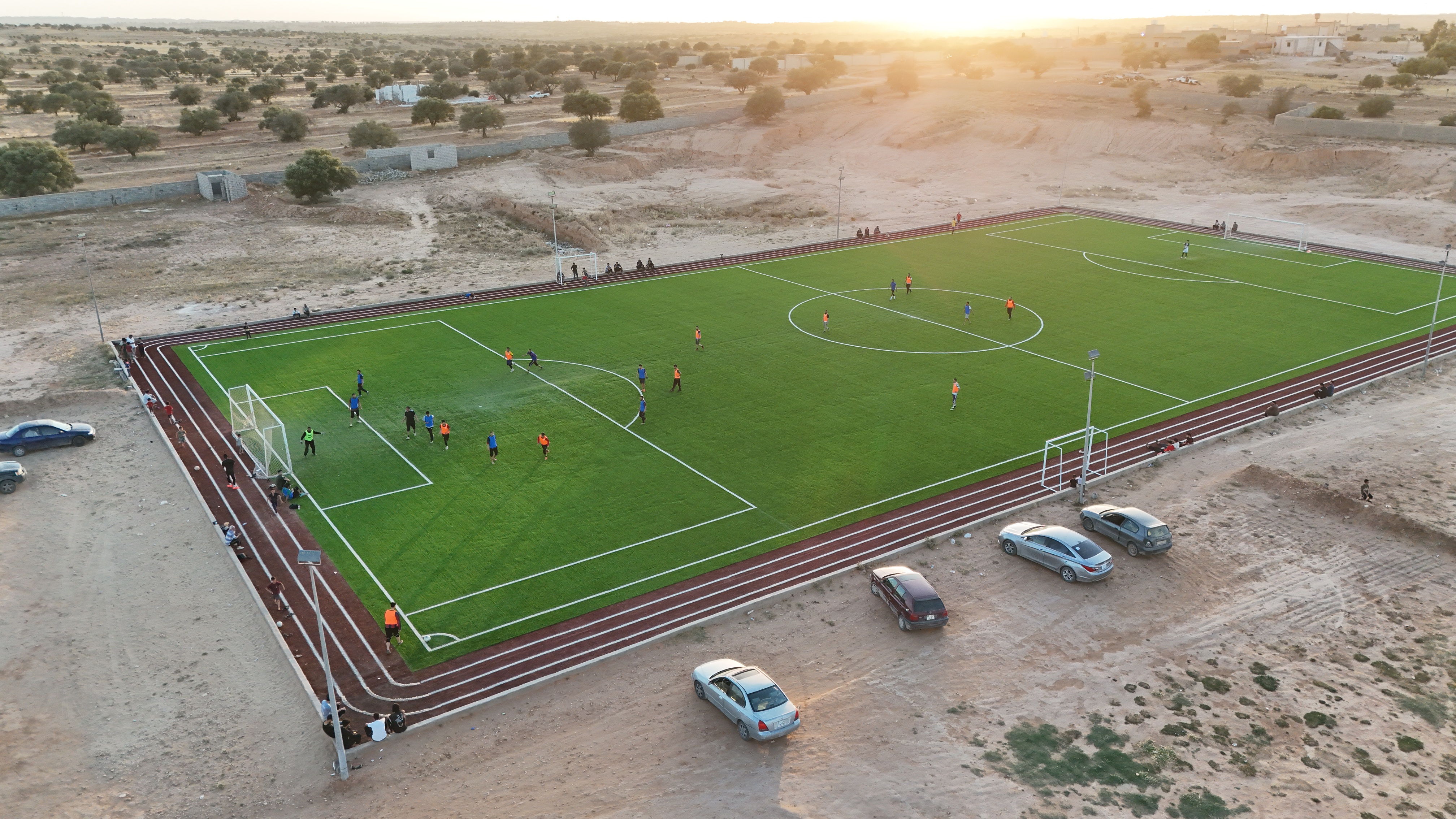 Aerial view of a soccer field with players and parked cars at sunset.