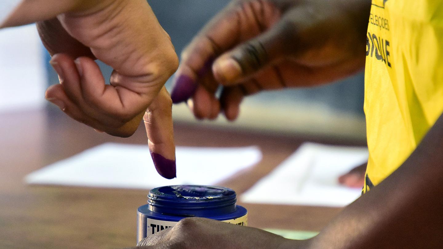 Image of a person dipping their finger into the voting ink box and a UNDP personnel guiding their hand.